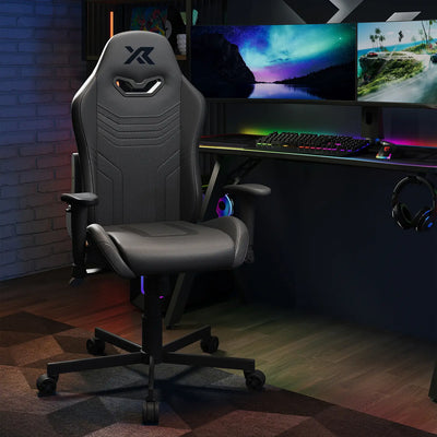 Agility Compact Gaming chair in Carbon Black in a dark room with a desk setup featuring monitors and RGB lighting.