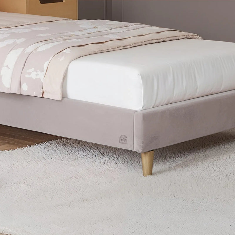 Beige upholstered bed with white mattress and light pink blanket on a carpeted floor.