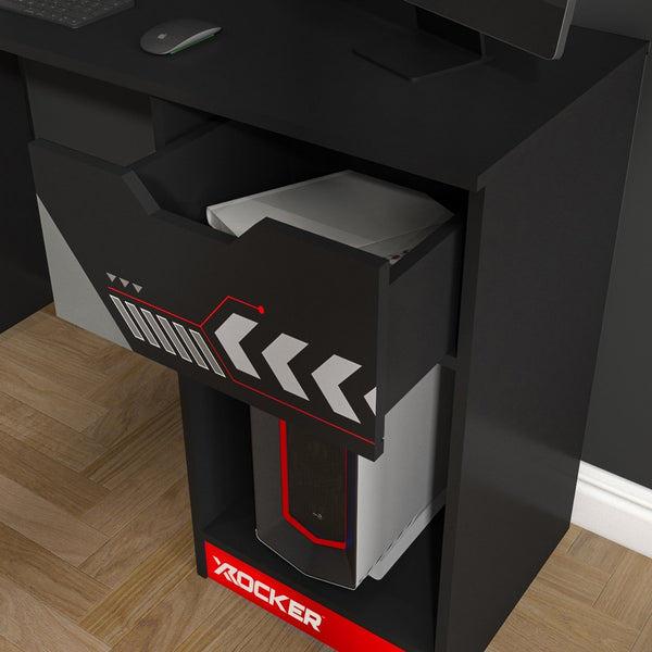 Black gaming desk with open drawer displaying a computer case, on a wooden floor.