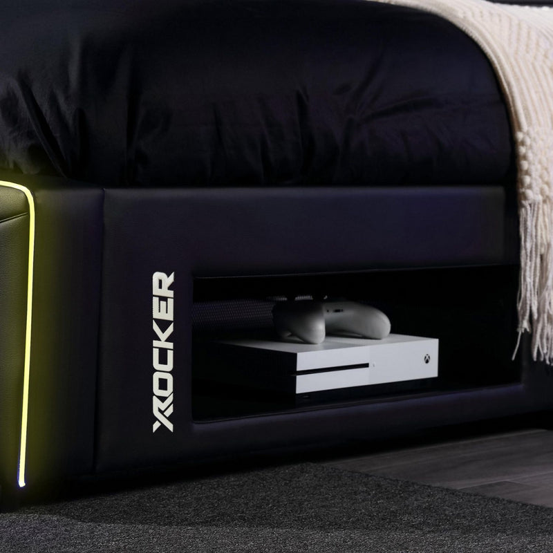 Black gaming bed with X rocker logo and gaming console inside, on a dark floor.