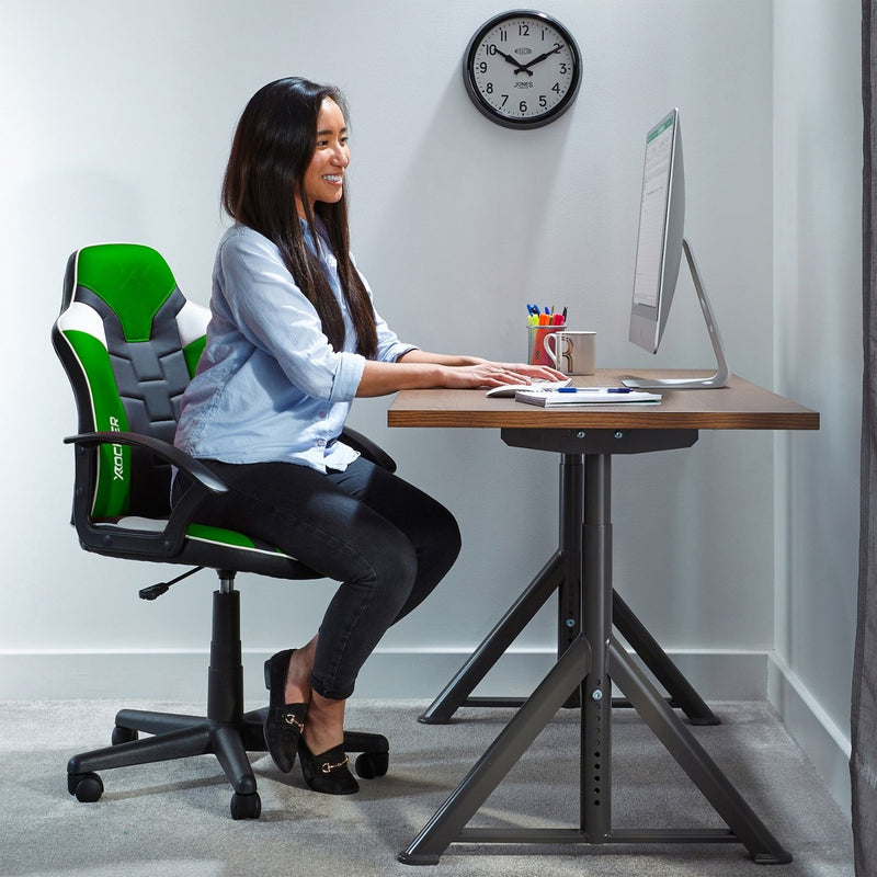 Woman sitting at a desk with a green and black gaming chair, using a computer.