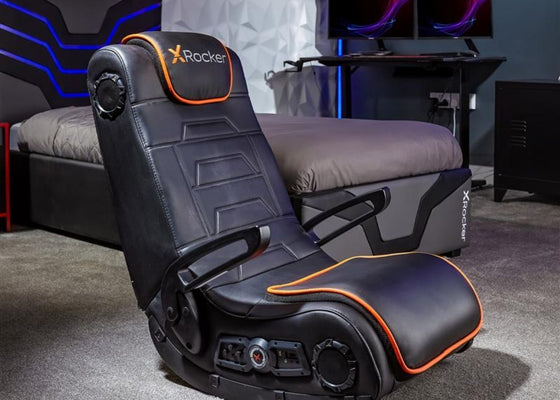 Black gaming chair with orange accents in a room setup with a desk and monitors.