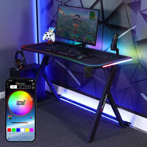 An RGB gaming desk with a smartphone app interface displayed, showing customizable lighting colors and patterns. The desk features a black color, metal and plastic construction, with a monitor, keyboard, and a gaming mouse set up on it.