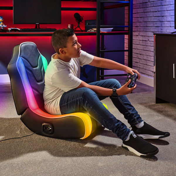 A child seated on a black gaming chair with multicolored LED lights, playing a video game with a controller.