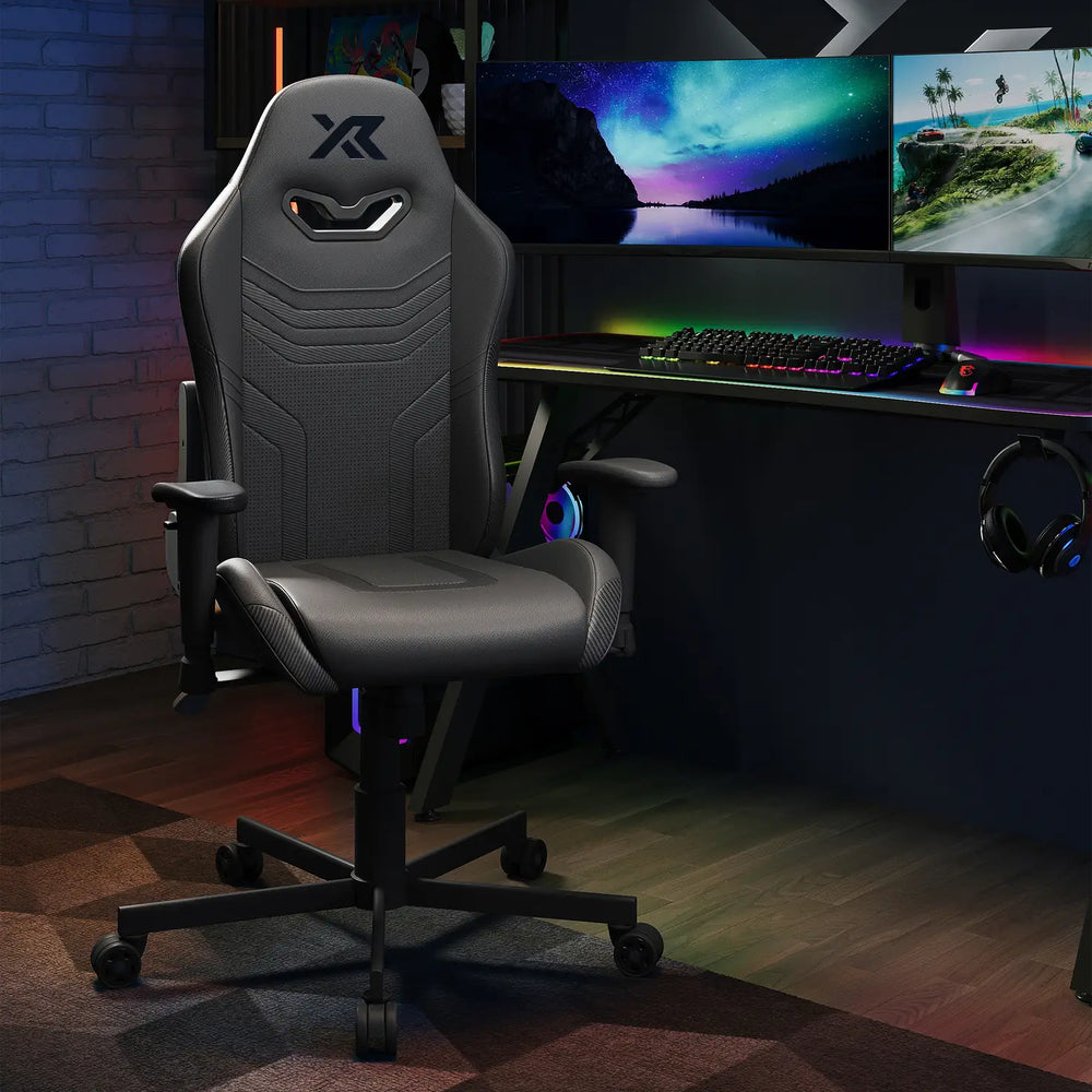 Agility Compact Gaming chair in Carbon Black in a dark room with a desk setup featuring monitors and RGB lighting.