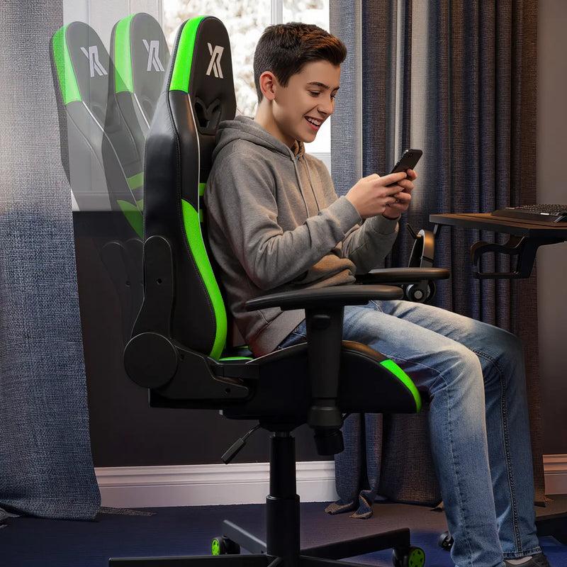 Person sitting on a black and green gaming chair using a smartphone in a room with curtains.