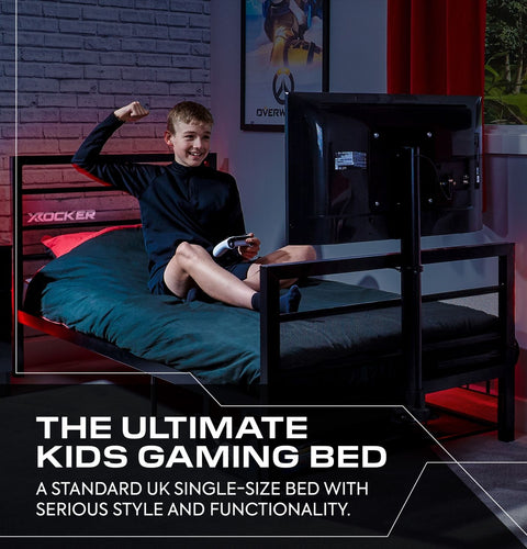 Child sitting on a gaming bed with a TV, surrounded by gaming equipment in a room with brick walls.