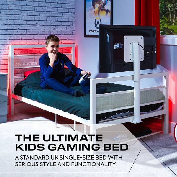Child sitting on a gaming bed with a TV stand, surrounded by gaming equipment in a room with red and white decor.