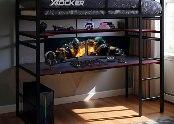 Black X rocker gaming desk with two monitors, keyboard, and mouse in a room.