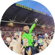 Ben Foster in green shirt holding a smartphone with a crowd and stadium in the background