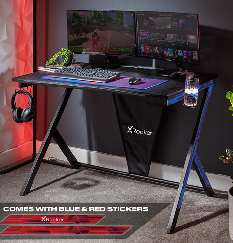 Gaming desk with X rocker branding, two monitors, keyboard, and mouse in a room setting.