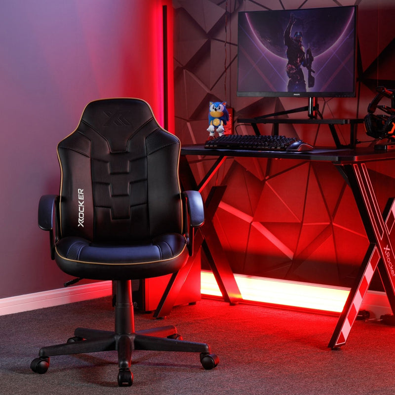 Gaming setup with a black X rocker chair, desk, and computer setup in a room with red lighting.