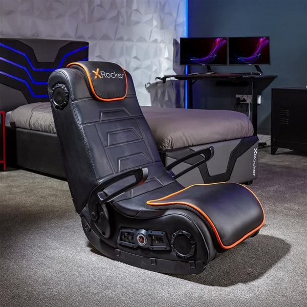 Black gaming chair with orange accents in a room setup with a desk and monitors.