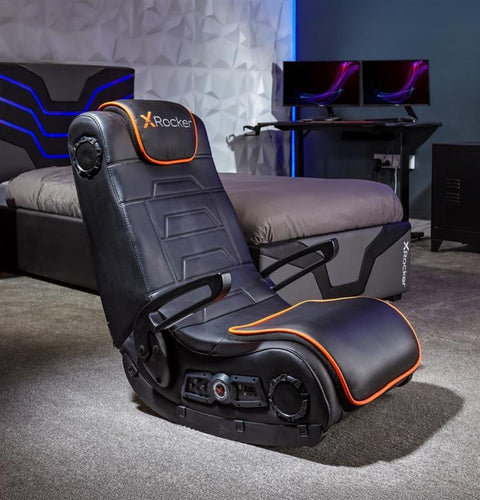 Black gaming chair with orange accents in a room setup with a desk and monitors.
