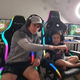 Person and child sitting in colorful gaming chairs in a room with computer monitors.