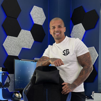 Man in a white t-shirt with a logo sitting in front of a computer setup with acoustic foam panels on the wall.