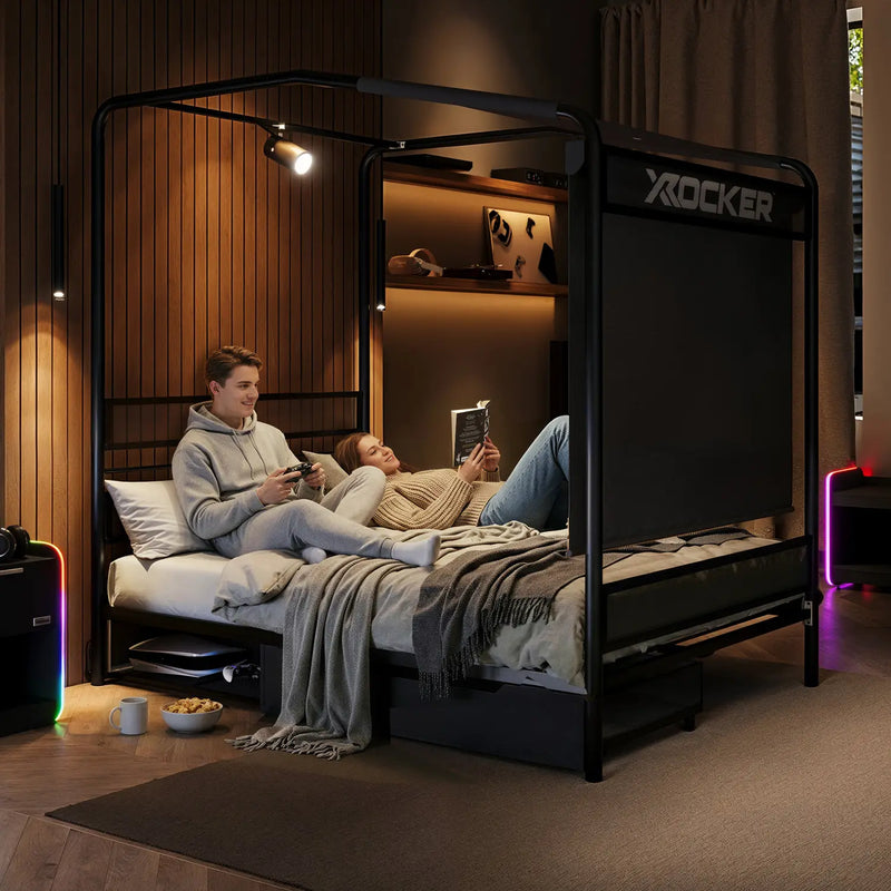 Two people lying on a black 'X rocker' gaming bed in a dimly lit room.
